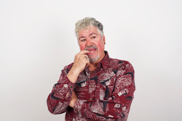 Dreamy male with thoughtful expression, looks to the camera, keeps hand near face, bitting a finger thinks about something pleasant, poses against gray wall.