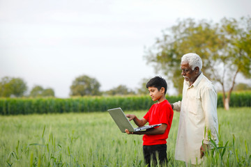 indian farmer with his grand son at wheat field, using laptop