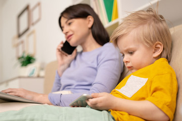 Cute blond little boy with smartphone watching online cartoon by his mother