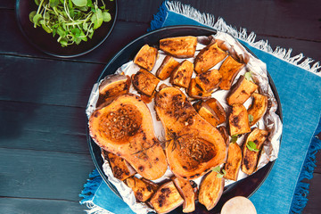 Top view baked roasted grilled whole orange pumpkin butternut squash halfs and sweet potato slides with herbs on the baking sheet on blue napkin and black wooden table. Vegetarian and vegan food diet.