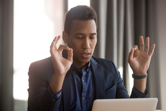 Mixed race businessman sitting at desk workplace closed eyes folded fingers mudra symbolic gesture relief stressful situations, control negative emotions, improve self-control, reduce fatigue concept