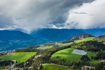 Aerial view of farm of solar batteries on green slopes of the mountains of Italy, Trentino, huge clouds over a valley, roofs of houses of settlements, green meadows, a clear energy, energy of the sun