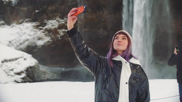 beautiful young woman near a waterfall in iceland, takes pictures on the phone, rejoices and takes a selfie