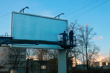 two workers on a hydraulic lift platform stick a poster on a billboard
