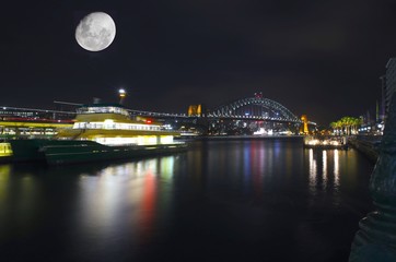 Fototapeta na wymiar Sydney harbour illuminated by the moon and circular quay with vibrant colourful lights at midnight in NSW Australia