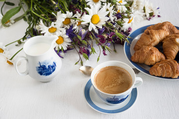 Summer morning breakfast. Cup of coffee, hot croissants and bunch of wild flowers on white wooden background