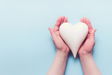 Obraz premium Female hands holding white porcelein heart on blue backgound. Photo with copy blank space.