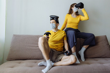 Fototapeta na wymiar A young couple stays at home during the epidemic and plays games. The guy and the girl are dressed the same. People are immersed in a virtual 3D game. The couple is sitting on sofa in the living room