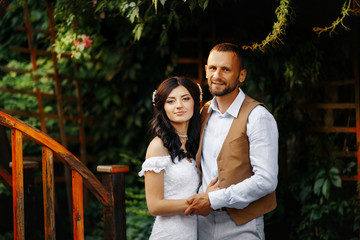 A happy, stylish couple in love posing on the bridge under a blossoming bush on their wedding day. A romantic colourful bright portrait of the bride and groom in a long white dress with a trail.