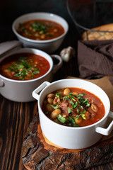 Bean soup with meat and vegetables served on a rustic board and wooden table with bread and garlic. Traditional Balkan soup Pasulj (Grah). Close-up, selective focus