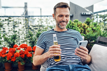 Smiling man relaxing on terrace with beer using smartphone