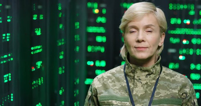 Portrait of beautiful serious Caucasian military female officer in camouflage looking at camera in dark monitoring room. Digital analytic center concept. Close up of US army woman among servers.