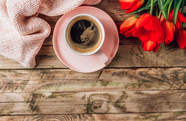 A pink cup of coffee, knitted plaid and real tulip flowers on a rustic wooden background. Top view. Flat lay. Female workplace with cozy breakfast