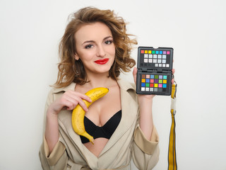 Sexy woman with red lips and curly hairdo in short coat and black bra playfully posing with banana and color calibration target passport on white background