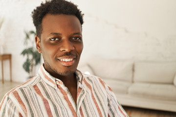 Close up image of cool cheerful young dark skinned man with Afro hairstyle sitting in stylish living room interior, relaxing at home, looking at camera with broad happy smile. People and lifestyle