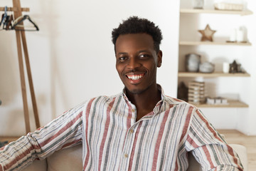 Obraz premium Indoor image of joyful positive young dark skinned man with stylish afro hairstyle posing against cozy living room interior background looking at camera with happy smile, feeling relaxed and carefree
