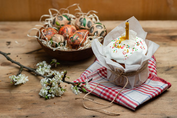 Easter cake with candle in parchment paper on a red checkered tablecloth and easter eggs in clay plate on wooden board with a flowering branch on a wooden background