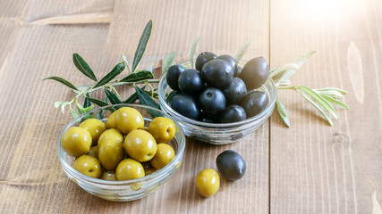 Black and green olives in glass bowls, olive tree branch on the wooden table in sunlight closeup....