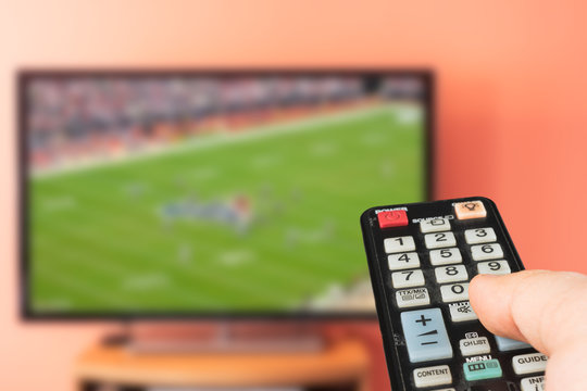 Caucasian man watching american football game on TV at home. Changing channels and adjusting volume with television remote control. 