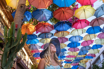 blond young  woman in dark blue hat and street of colorful umbrellas above her in north part of Nicosia the capital city of Cyprus - obrazy, fototapety, plakaty