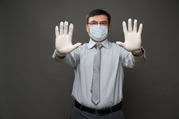 a man dressed as a businessman showing stop with the palm of his hand, gray background, medical face mask and protective gloves, glasses, shirt and tie - concept of quarantine and antivirus protection