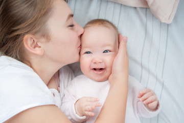 Mother kissing her infant baby in bed. Happy mother kisses baby lying on the bed. Beautiful smiling girl lying on her back. Top view. Flat Lay.
