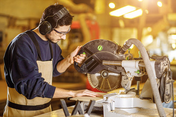confident young carpenter work with mitre saw in factory, stand standing behind a workbench wearing uniform. industry, handicraft, carpentry concept