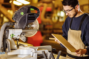 skilled young woodworker in safety gear going to cut piece of wood with of mitre saw or circular saw while working alone in woodworking studio