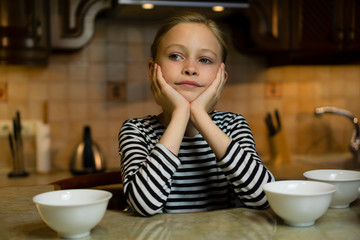 Obraz na płótnie Canvas Caucasian blond child girl in a striped dress does not know what to cook sitting at cosy home kitchen table with empty white plates bowls