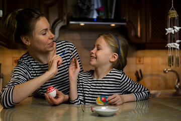 Beautiful caucasian mother and child daughter in striped dresses having fun paint easter eggs sitting at table at home cosy wooden kitchen