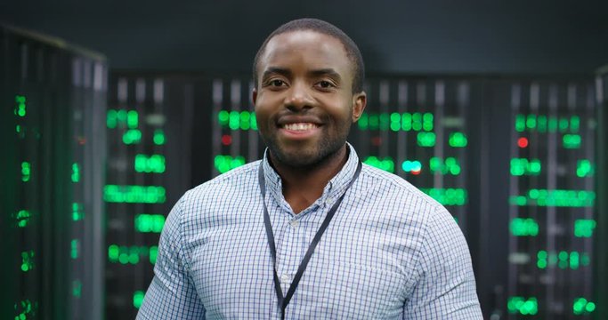 Portrait of young handsome African American man smiling cheerfully too camera in database center full of computers and servers. Close up of male worker of server room. Cybersecurity world.