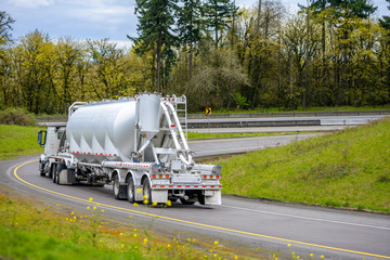 Big rig day cab semi truck with bulk semi trailer turning on the highway entrance with green trees on the sides