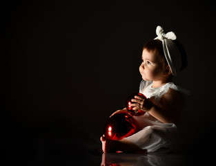 Little female in white headband and dress, barefoot. She holding two red balls, sitting on floor. Twilight, black background.
