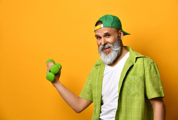 Grandpa in green cap and shirt, white t-shirt, bracelet. Holding two dumbbells and pointing at them, posing on orange background