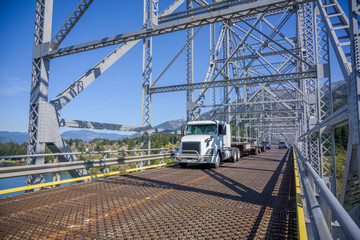 Naklejka premium Day cab powerful big rig white semi truck with empty step down semi trailer driving on the silver truss bridge across Columbia River