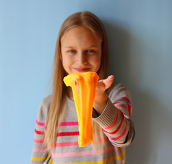 A blonde girl playing with orange handmade slime on blue background. The focus is on slime