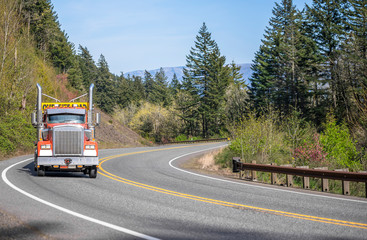 Orange big rig semi truck with oversized load sign on the roof running uphill on the winding road