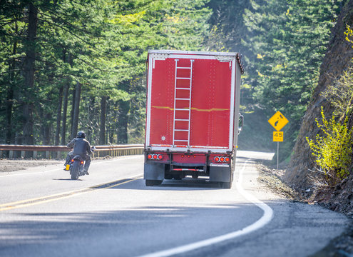 Motorcyclist overtaking a big rig semi truck with red bulk semi trailer on the winding mountain road in prohibited no-overtaking zone without a secure road view