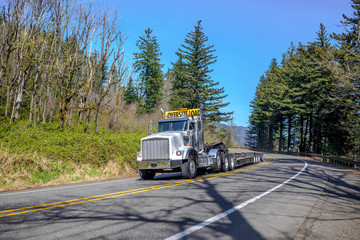 Fototapeta na wymiar Powerful big rig white semi truck with oversize load sign on the roof transporting empty step down semi trailer for carry heavy-duty cargo running on the road with forest on the hill