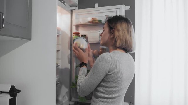 Woman opens refrigerator door in kitchen at home and takes container with food