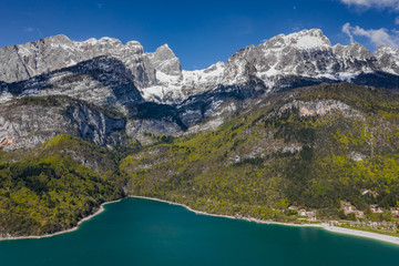 Naklejka na ściany i meble The Improbable aerial landscape of village Molveno, Italy, azure water of lake, empty beach, snow covered mountains Dolomites on background, roof top of chalet, sunny weather, a piers, coastline, 