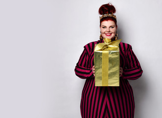Fototapeta premium Overweight ginger female in striped dress, crown and earrings. Holding golden gift box, excited, posing isolated on white