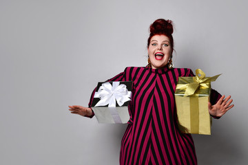Obese lady in striped dress and earrings. Holding golden and silver gift boxes, overjoyed, posing on white photo background