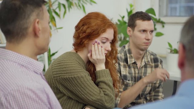 Tilt up shot of red-haired depressed young woman telling her sad story to other patients sitting in circle during group therapy session