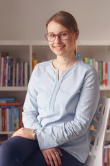 Front view portrait of beautiful young adult caucasian woman female girl sitting in the chair at home or office in front of the bookshelf looking to the camera wearing eyeglasses shirt in day smiling