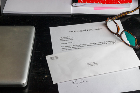 A notice of furlough lays on a table with an envelope, computer, notebook, and reading glasses.