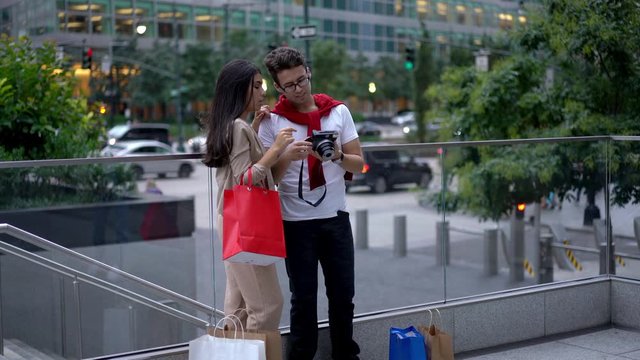 Happy man and woman dressed in stylish wear taking pictures on old fashioned equipment after shopping time in downtown, cheerful couple in love smiling at camera while taking selfie
