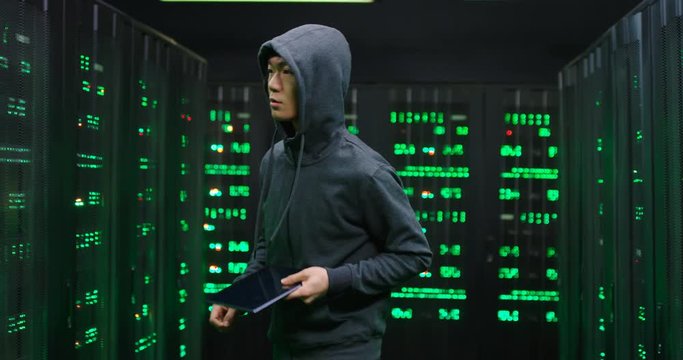 Male Asian guy data thief in dark hood stealing in the server room with tablet device in hands. Digital spy man hacking database. Male hacker commiting cyber crime.