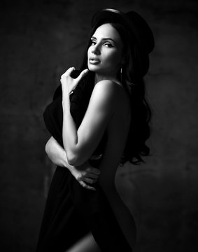 Young beautiful stylish brunette naked woman in hat standing and holding black clothing in hands. Black and white image