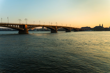 Die Theodor-Heuss-Brücke ragt umgeben vom wunderschön blauen Rhein im Sonnenuntergang in die mainzer Skyline